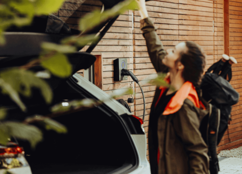 Eine Frau mit Rucksack schließt den Kofferraum eines Elektrofahrzeuges, im Hintergrund ist noch das Kabel der Elektroladesäule zu erkennen.
