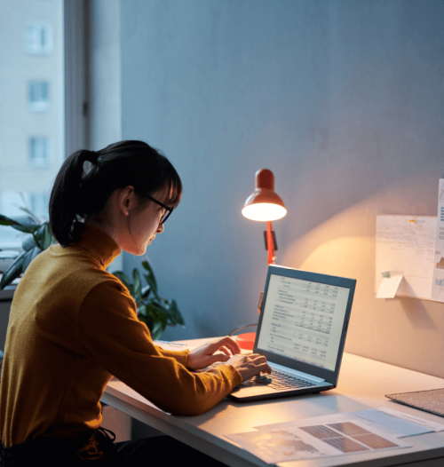 Eine Frau sitzt am Schreibtisch vor ihrem Laptop, eine Schreibtischlampe leuchtet.