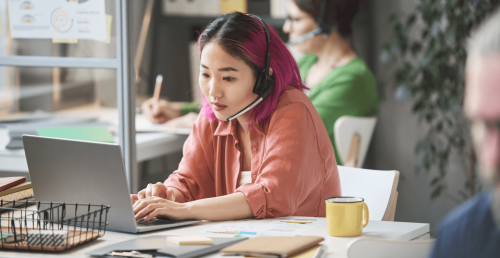Beraterin mit Headset unterstützt Kund:innen am Laptop in modernem Büro mit Unterlagen und Diagrammen.
