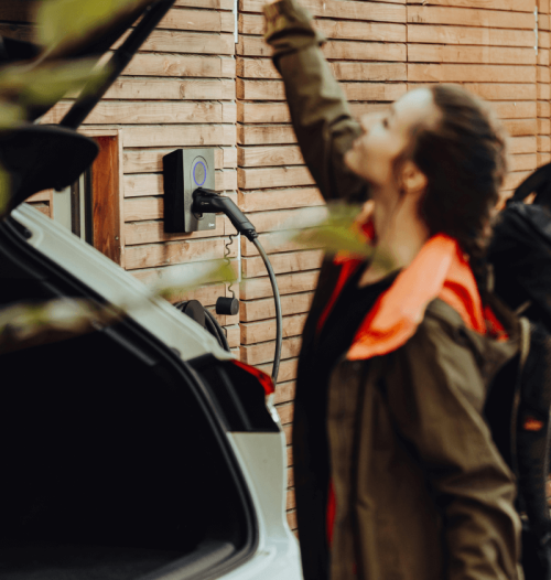 Eine Frau mit Rucksack schließt den Kofferraum eines Elektrofahrzeuges, im Hintergrund ist noch das Kabel der Elektroladesäule zu erkennen.