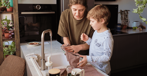 Mutter zeigt ihrem Sohn hilfsbereit wie man den Abwasch vornimmt es läuft noch an der Küchenzeile der Wasserhahn.