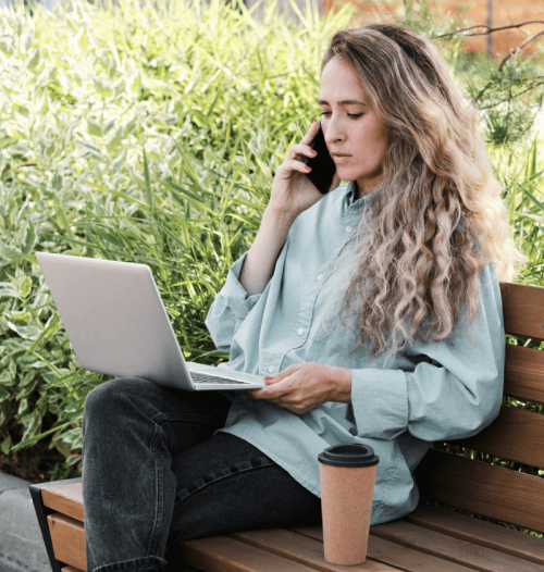 Eine Frau hat es sich gemütlich auf einer Parkbank gemacht, telefoniert und schaut in ihren Laptop