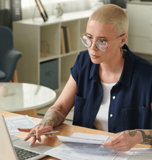 Eine Frau mit Brille prüft konzentriert Dokumente am Laptop.