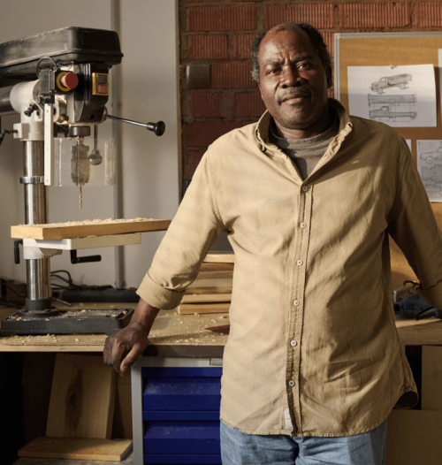 Handwerker steht in Werkstatt neben Ständerbohrmaschine, mit Holzspänen und technischen Zeichnungen im Hintergrund.